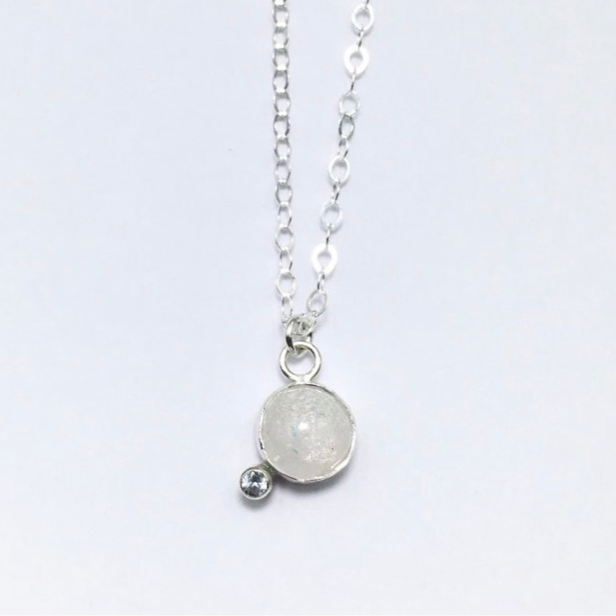 A silver necklace with a round milk stone pendant and a smaller round birthstone pendant attached to the milk stone.